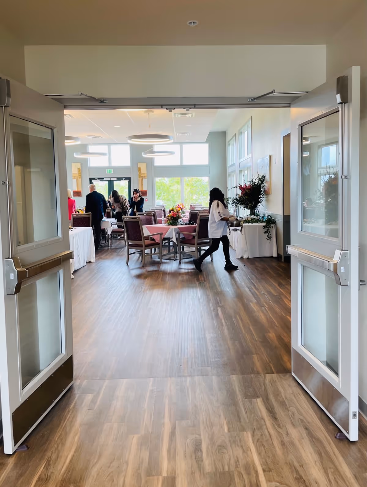 View through open double doors into a bright dining room with wooden floors, tables covered with pink tablecloths, chairs, and floral centerpieces. Several people are standing and sitting at the tables near large windows letting in natural light.