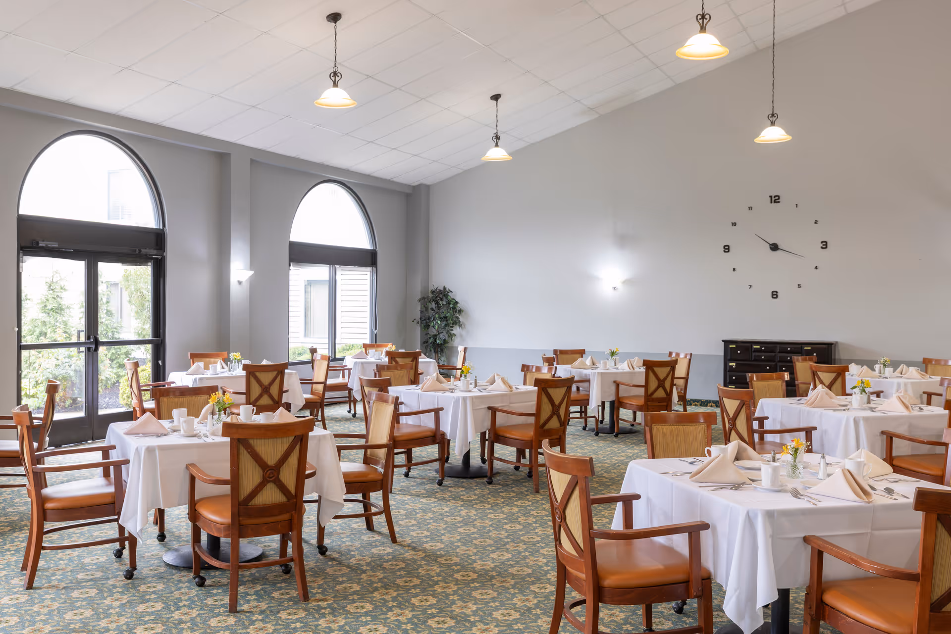 Bright dining room with multiple round tables set with white tablecloths and wooden chairs under pendant lights.