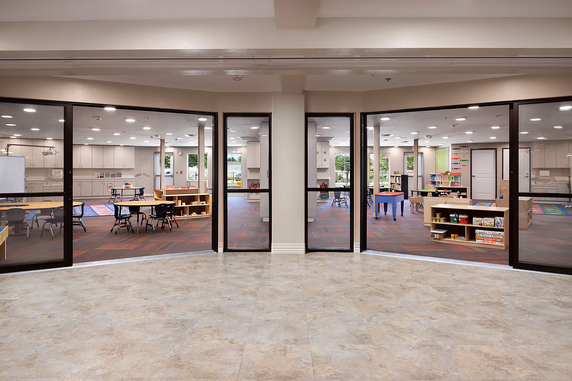 Wide interior view through glass walls into an activity/playroom with small tables, chairs, and shelving for toys and supplies.