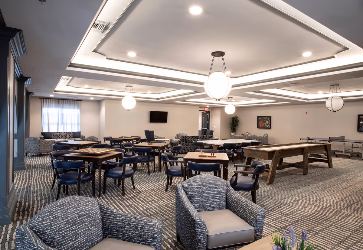 A spacious common area in a senior living facility with multiple tables and chairs arranged for games and socializing. The room features comfortable armchairs, a shuffleboard table, a ping pong table, and a wall-mounted TV. The ceiling has recessed lighting and large round pendant lights, with neutral-colored walls and patterned carpet.