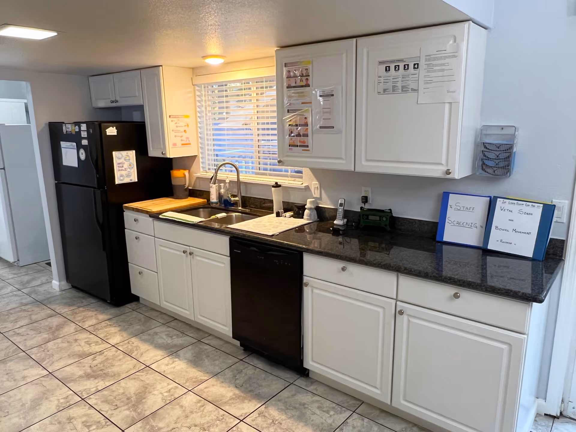 White galley-style kitchen with black granite countertops, a sink under a window, black refrigerator and dishwasher, and tiled floor.