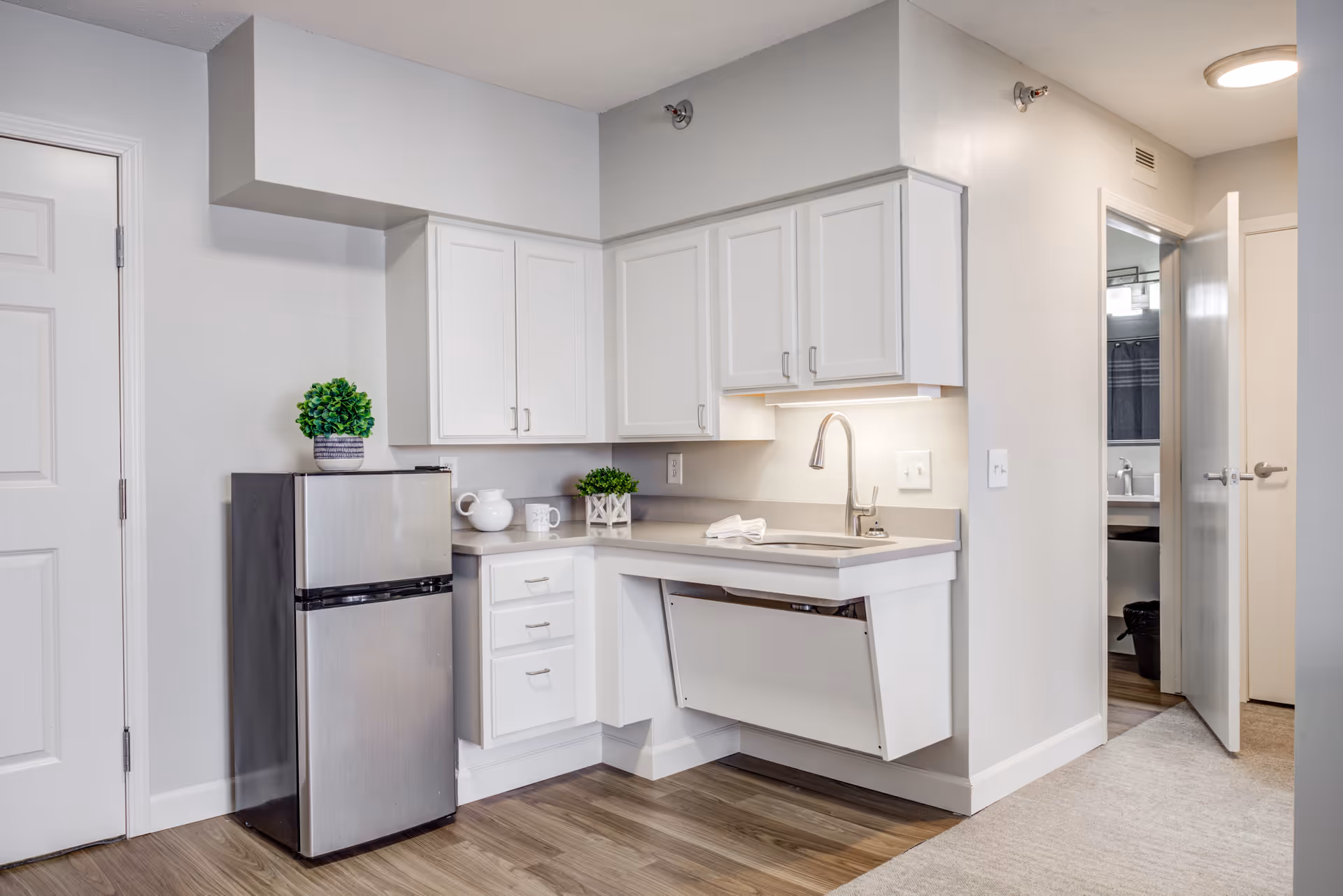 A small kitchen area with white cabinets, a stainless steel mini refrigerator, a sink with a modern faucet, and a countertop with decorative plants and a white pitcher. The floor is a combination of wood and carpet, and there is an open door leading to a bathroom visible in the background.