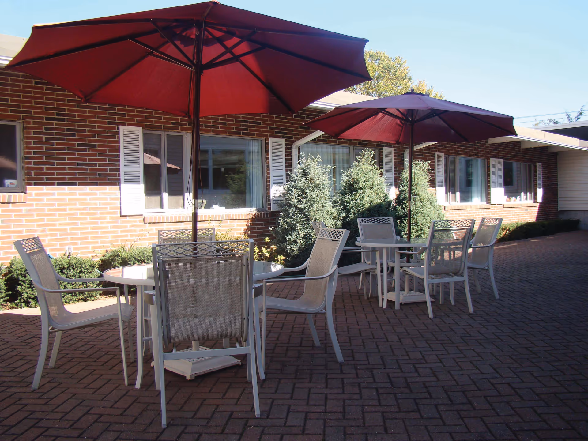 Outdoor patio area with two round tables, each surrounded by four white chairs and shaded by large red umbrellas. The patio is paved with brick and is adjacent to a brick building with white window shutters and some greenery.