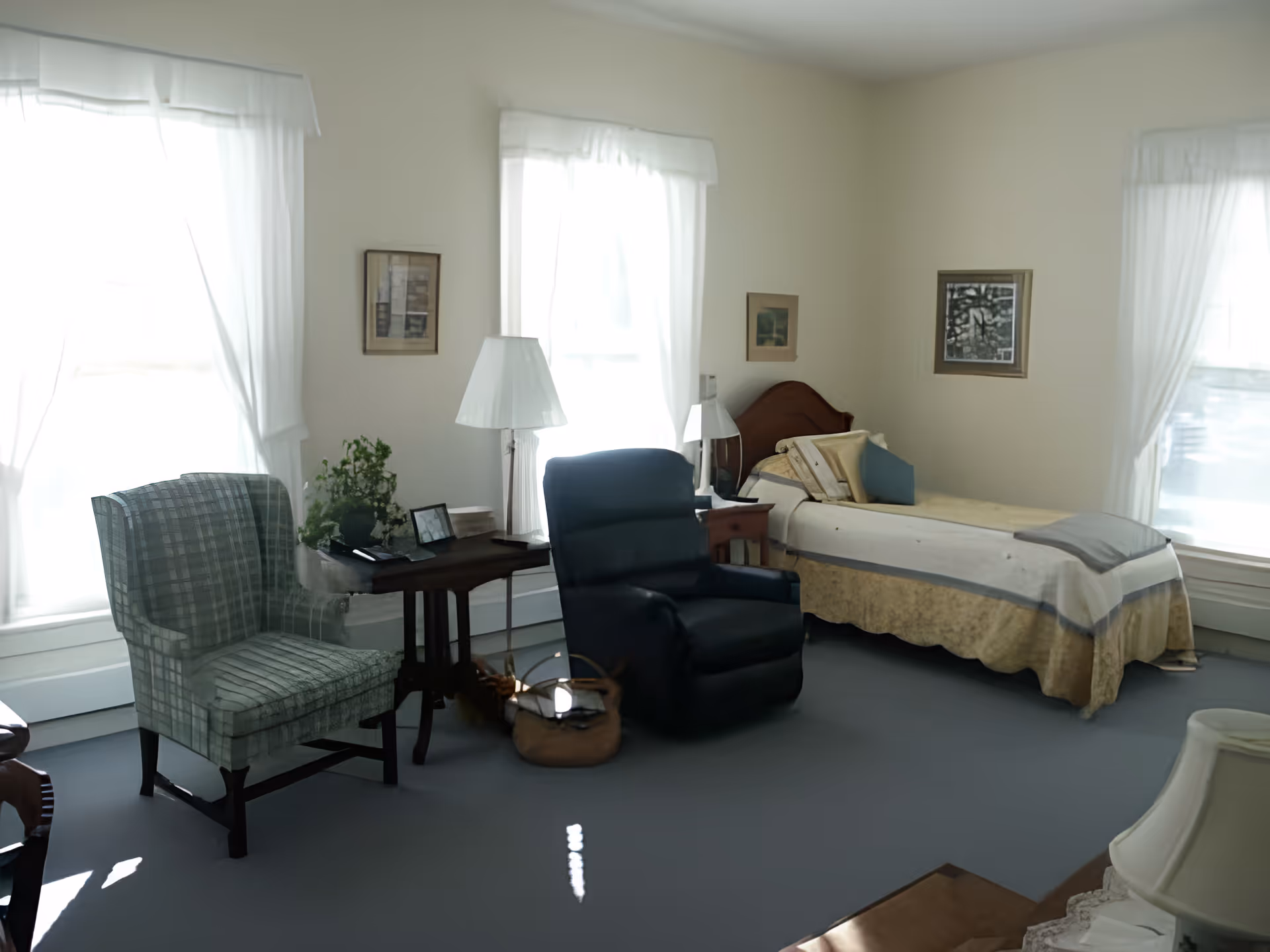A bright and cozy bedroom with a single bed covered in a beige and gray quilt, a dark blue recliner chair, a green plaid armchair, a wooden side table with a lamp and a plant, and three large windows with white curtains letting in natural light. The walls are decorated with framed pictures.
