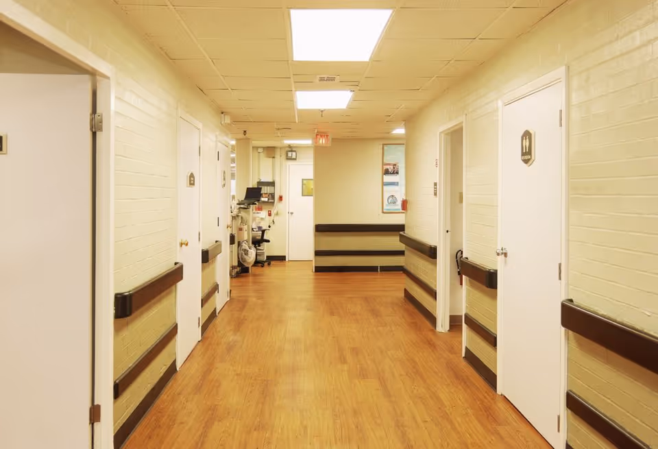 Well-lit interior corridor with wood flooring, wall handrails and several closed doors leading to rooms.