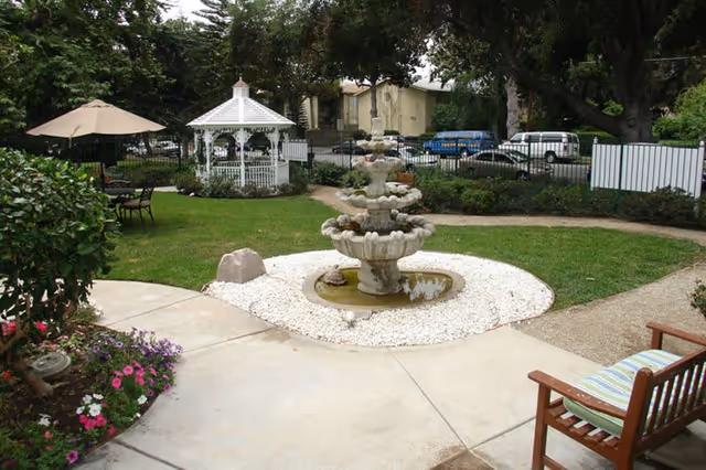 Outdoor garden area with a multi-tiered stone fountain surrounded by white pebbles, a white gazebo, patio tables with umbrellas, green grass, trees, and a wooden bench with a striped cushion.