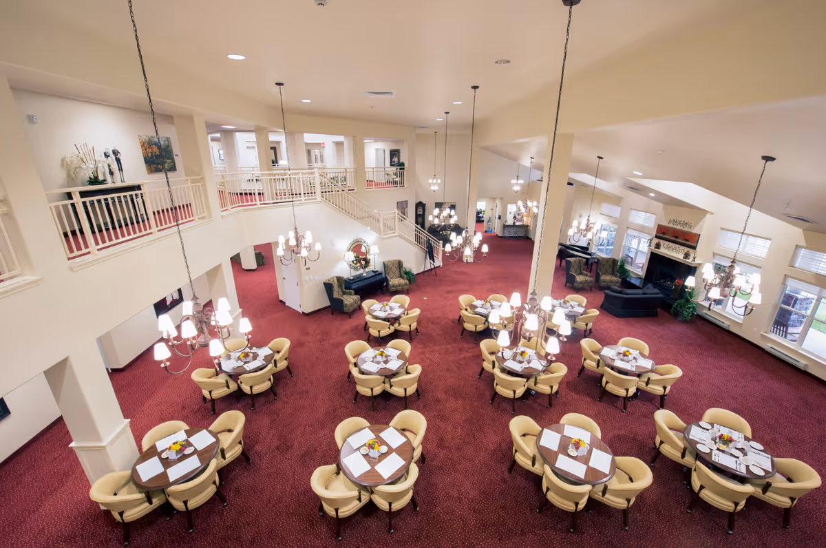 Large open dining room with multiple round tables, yellow chairs, chandeliers, and red carpeting in a senior living facility.
