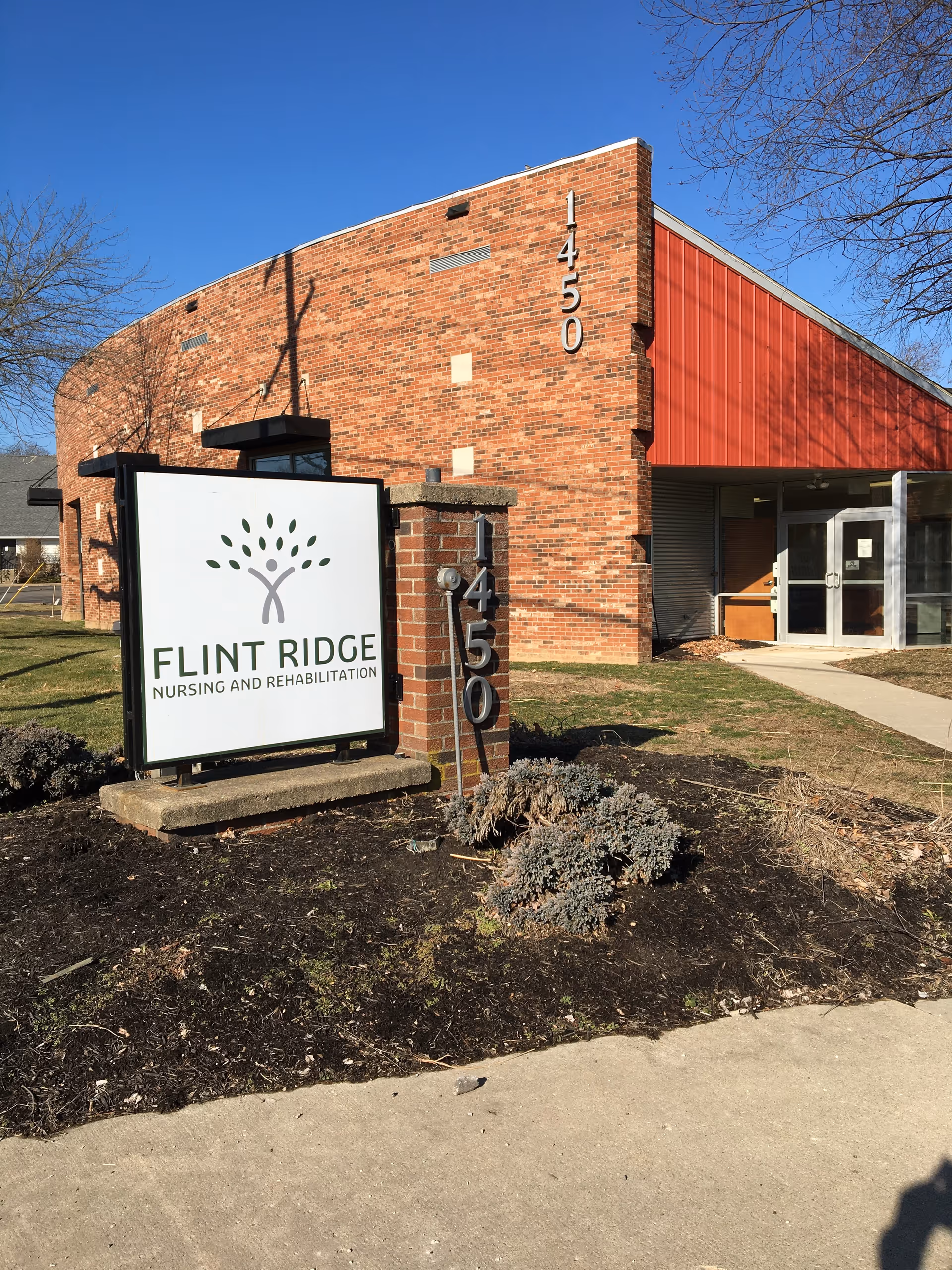 Exterior view of a brick building with the address number 1450 displayed vertically on the corner. In front of the building is a sign for Flint Ridge Nursing and Rehabilitation featuring a tree logo. The area around the sign has some landscaping with mulch and small shrubs. The sky is clear and blue.