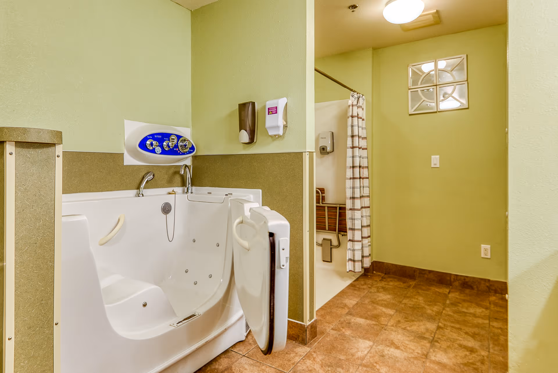 A bathroom with a walk-in bathtub featuring a door and handrails, a shower area with a curtain and a fold-down seat, light green walls, brown tiled floor, and a small window with a decorative grid.