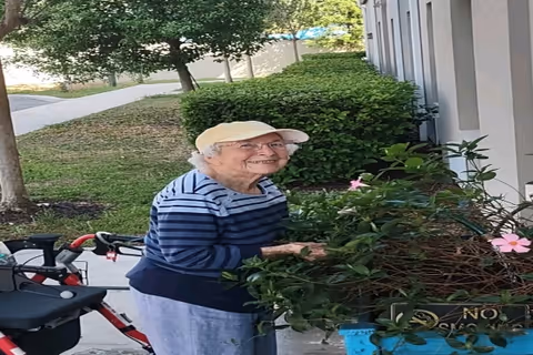 An elderly woman wearing a beige cap and a blue striped sweater is standing outdoors near a building, tending to a potted plant with pink flowers. There is a red and black walker nearby, and green bushes and trees are visible in the background.