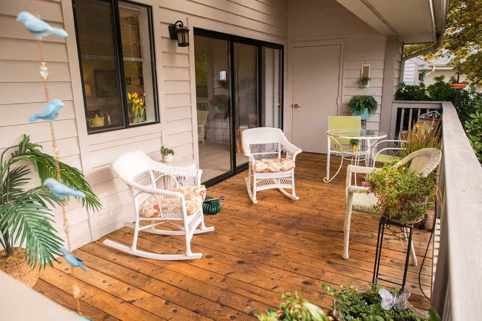 A wooden deck patio with white wicker rocking chairs and cushioned seats, a small glass-top table with a green chair, potted plants, and a sliding glass door leading inside a house.
