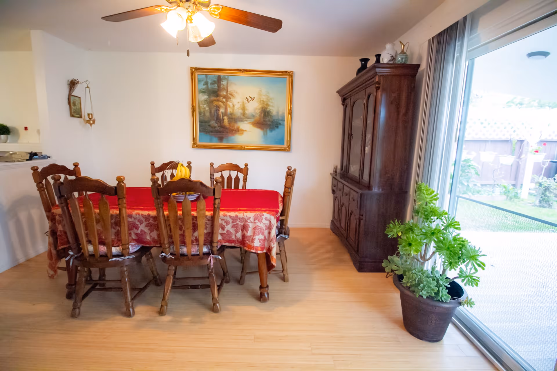 A dining room with a wooden table covered by a red and beige patterned tablecloth, surrounded by six wooden chairs. A ceiling fan with lights is above the table. On the wall, there is a framed painting of a nature scene with trees and birds. To the right, there is a large wooden cabinet with decorative items on top. A large potted plant is placed near a sliding glass door that leads to an outdoor area with a garden visible.