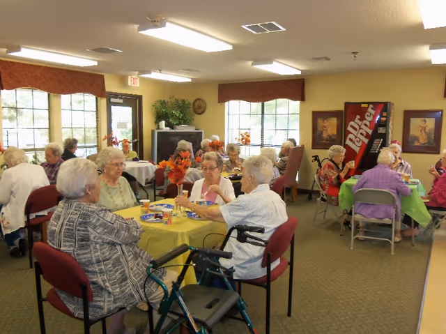 Elderly residents sit at tables with colorful tablecloths in a communal dining/activity room.