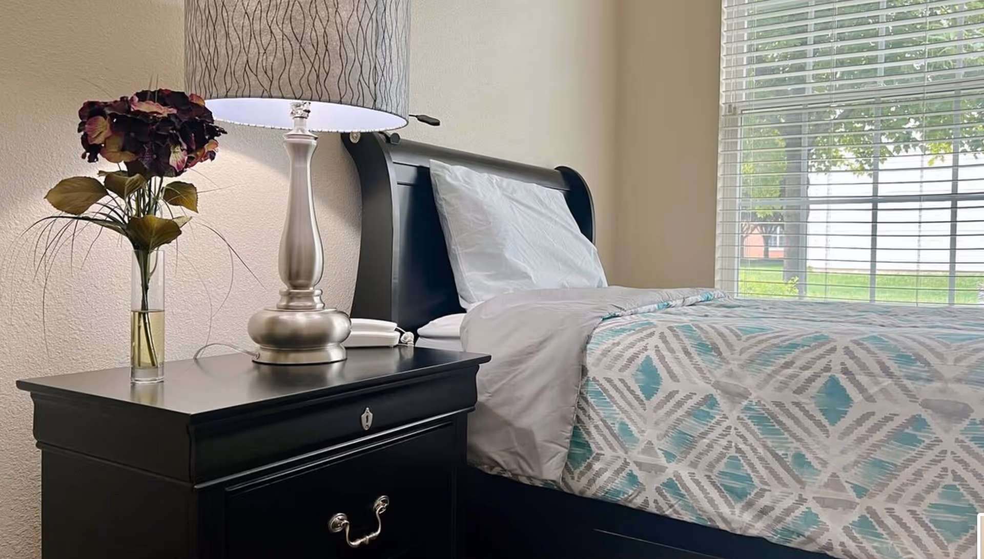 A neatly made bed with patterned bedding next to a black nightstand holding a lamp and a vase of flowers, beside a window with blinds.