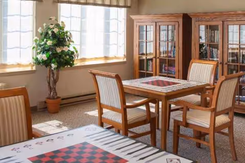 Sunlit senior living common area with wooden dining table, chairs, chessboard, bookshelf, plant near window.