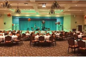 A large banquet hall with round tables covered in white tablecloths and set with chairs. The room features a stage at the front with a green curtain backdrop and decorative hanging lanterns. The ceiling has multiple chandeliers and recessed lighting.