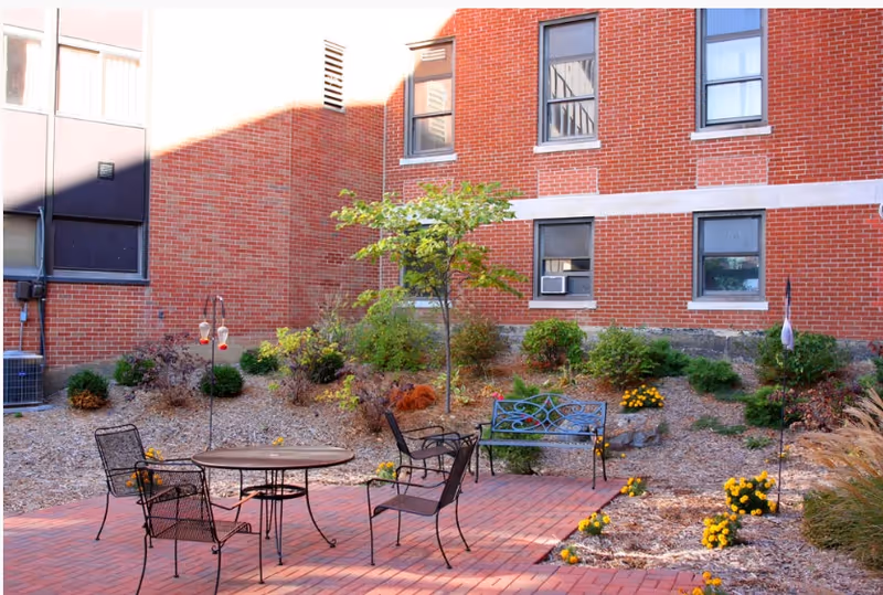 Outdoor courtyard area with a brick patio featuring a round metal table and four metal chairs. There is a metal bench with decorative backrest, surrounded by small bushes, flowers, and a young tree. The background shows a red brick building with several windows.