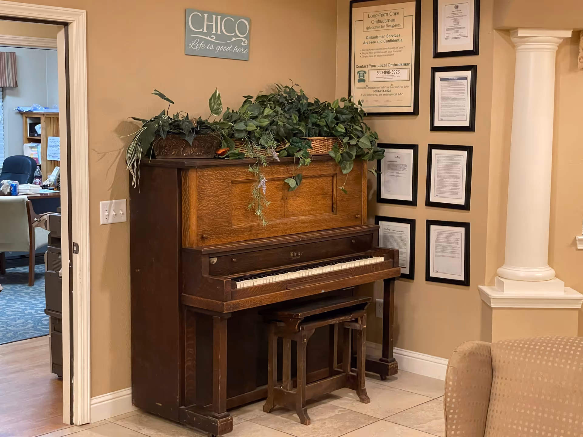 An upright wooden piano with a matching wooden bench in a beige-colored room. On top of the piano, there are several green leafy plants. Above the piano, a sign reads 'CHICO Life is good here'. To the right of the piano, there are framed documents and certificates hanging on the wall. A beige upholstered chair is partially visible in the foreground on the right side. An open doorway on the left leads to another room with office furniture and a blue carpet.