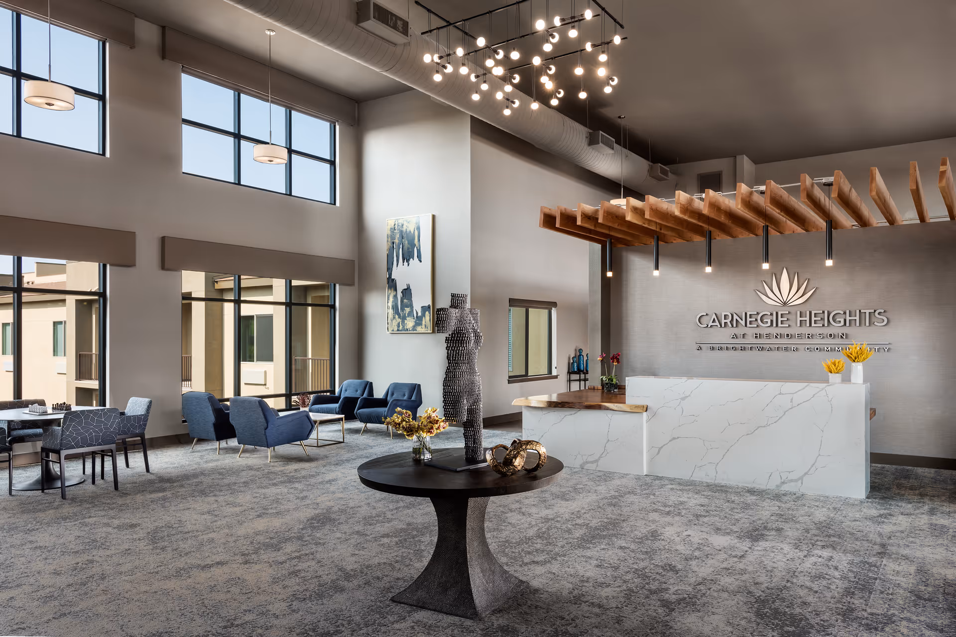 Spacious and modern reception area of Carnegie Heights at Henderson featuring a white marble reception desk with wooden accents, a decorative sculpture on a round table in the foreground, several blue armchairs and tables near large windows, and a contemporary chandelier hanging from the ceiling.
