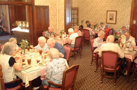 A group of elderly people sitting at multiple tables in a dining room, eating and socializing. The room has patterned wallpaper, carpeted floors, and wooden paneling. Tables are set with plates, glasses, and flowers in vases.