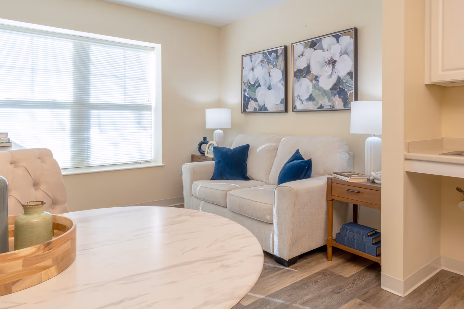 A cozy living room area with a beige sofa adorned with two dark blue throw pillows, two floral paintings on the wall above the sofa, two side tables each with a white lamp, a round marble table in the foreground with a wooden tray and a small green vase, and a window with blinds letting in natural light.