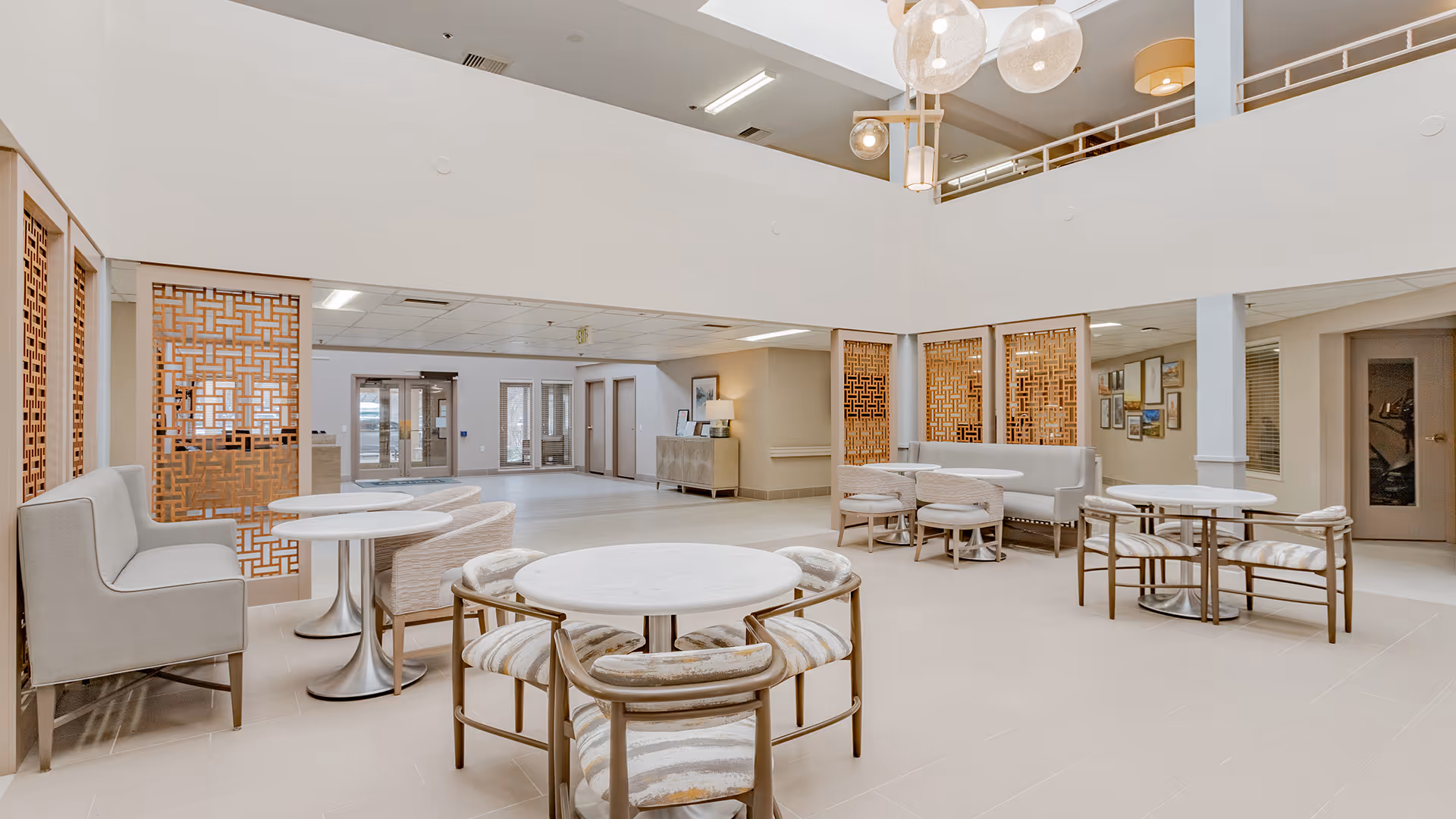 Bright and spacious common area in a senior living facility with round white tables, cushioned chairs, and bench seating. The room features a high ceiling with hanging light fixtures, decorative wooden lattice panels, and a neutral color palette. There is a second-floor balcony overlooking the area and doors leading to other rooms in the background.