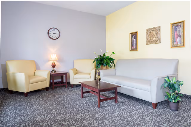 A cozy living room area with a beige sofa, two yellow armchairs, a wooden coffee table, a side table with a lamp, a wall clock, three framed artworks on a yellow wall, and two potted plants on a patterned carpet.