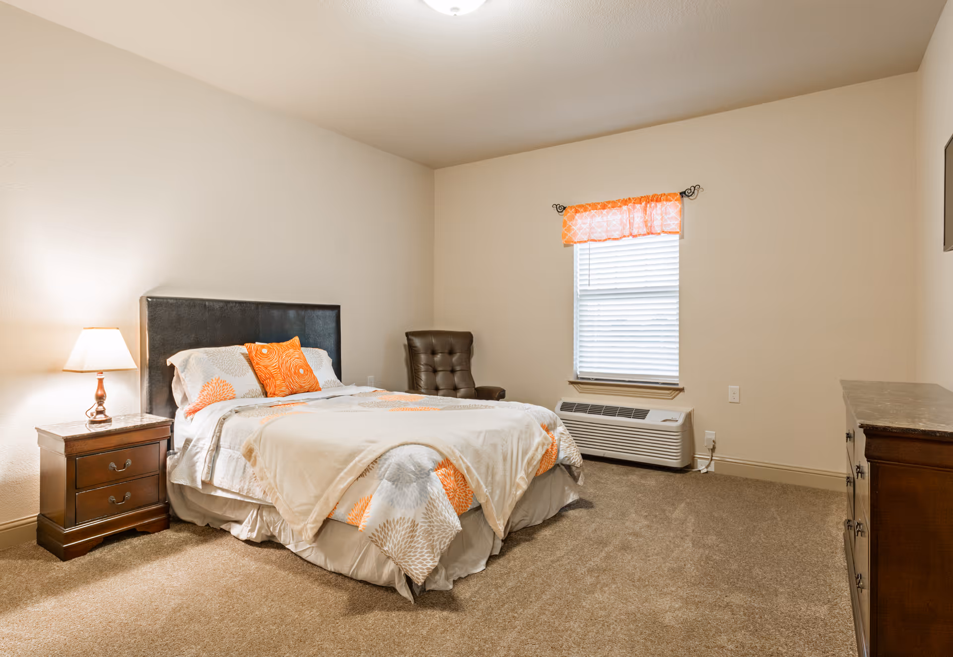 Furnished bedroom with a bed dressed in orange-accented bedding, a nightstand with lamp, an armchair, window with an orange valance, and a dresser.