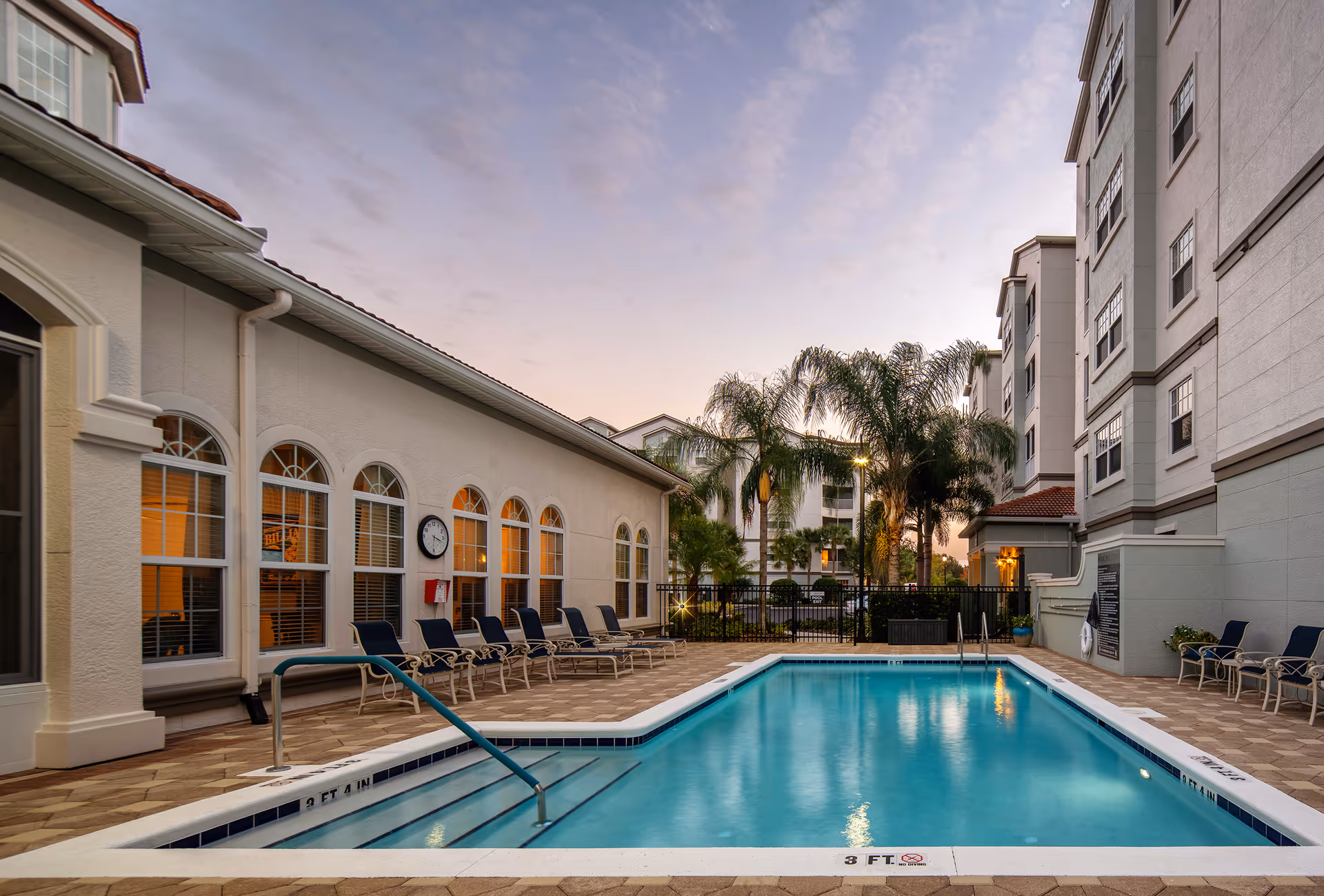 Outdoor swimming pool area at a senior living facility named Arietta, surrounded by lounge chairs and palm trees, with multi-story buildings in the background during dusk.