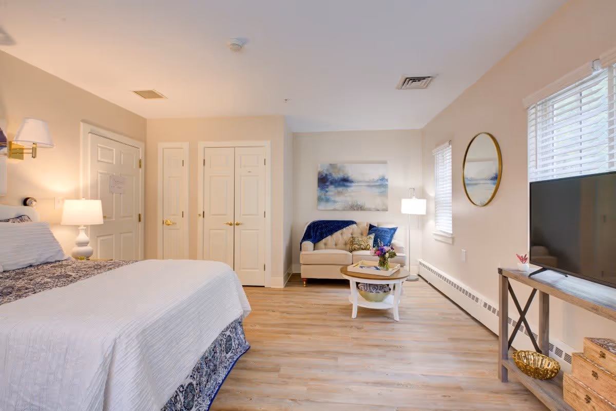 A bright and cozy bedroom with a large bed covered in white and patterned bedding. To the left, there is a bedside table with a lamp. In the background, there are two closed white doors and a small sitting area with a beige loveseat, a round wooden coffee table with flowers, and a floor lamp. On the right side, there is a flat-screen TV on a wooden stand, two windows with blinds, and a round mirror on the wall. The room has light-colored walls and wood flooring.