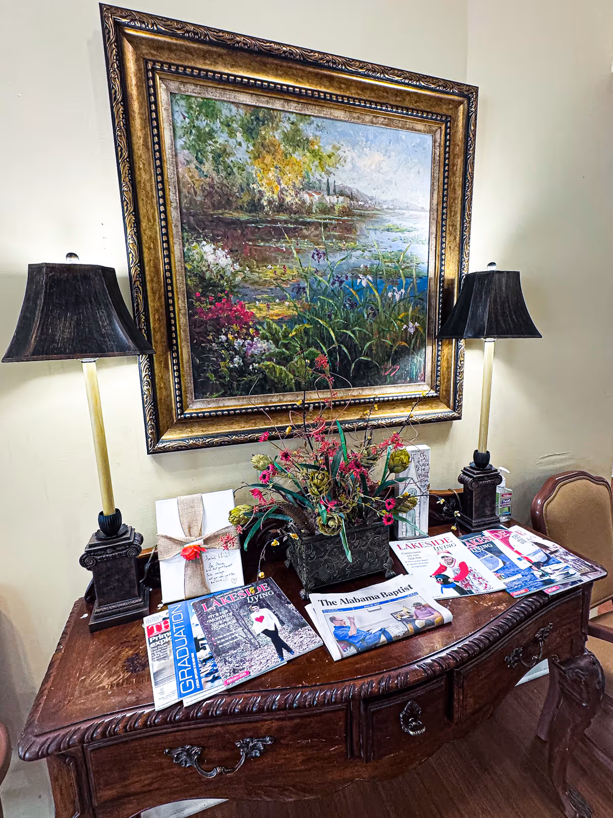 A wooden table with ornate carvings holds two black lamps with beige stands, a floral arrangement in a decorative vase, several magazines and newspapers, and a wrapped gift. Above the table hangs a large framed painting depicting a colorful landscape with a lake, trees, and flowers.