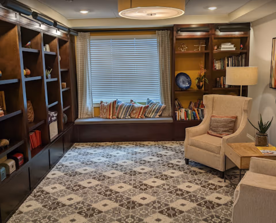 Cozy seating area with built-in bookshelves, a window seat with colorful pillows, two armchairs, side table and a patterned rug.