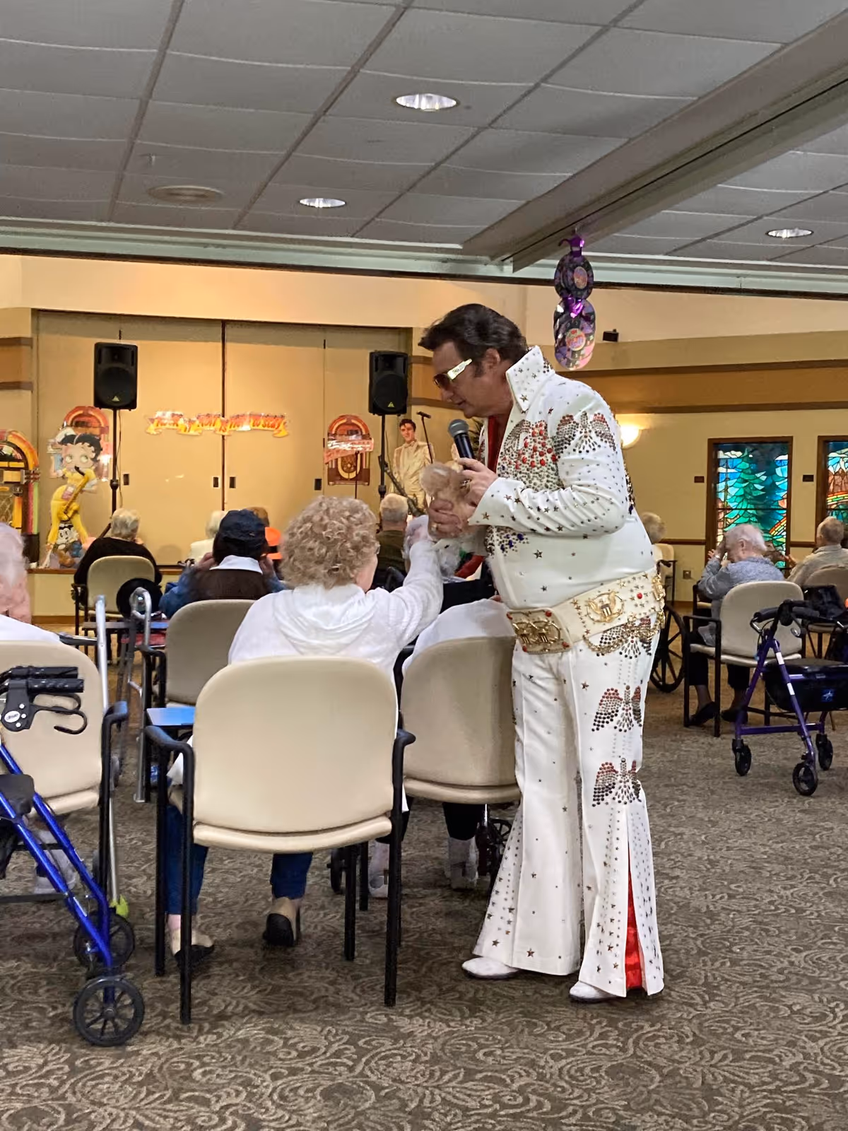 An Elvis Presley impersonator in a white rhinestone-studded jumpsuit is performing in front of a seated audience of elderly people in a senior care facility. The performer is holding a microphone and interacting with a woman in the front row. The room has beige walls, carpeted floors, and stained glass windows on the right side. Several walkers are visible near the seated audience.