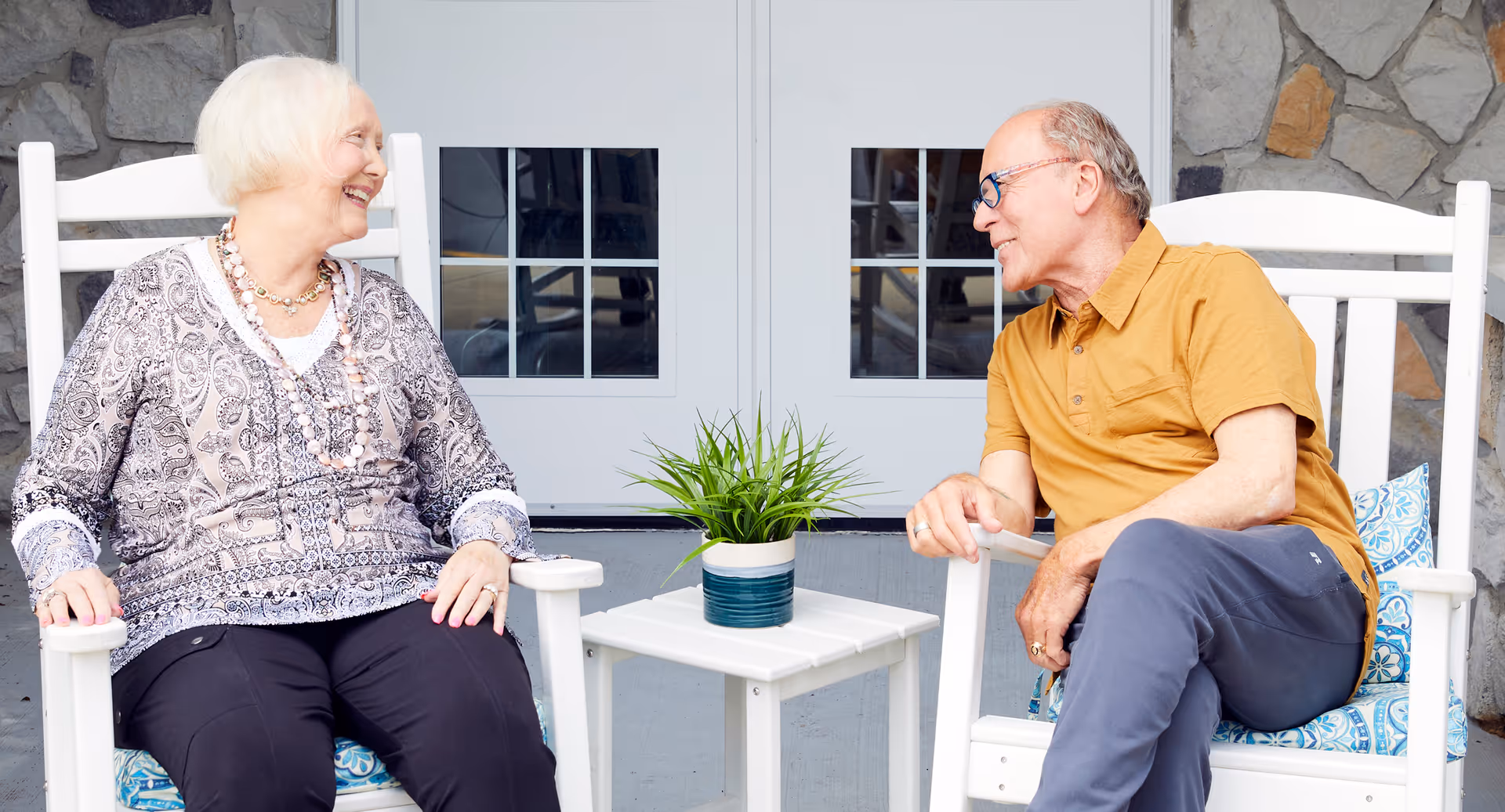 An elderly woman and man sitting on white rocking chairs on a porch, smiling and talking to each other with a small white table and a potted plant between them. The background shows a stone wall and white double doors with glass panes.