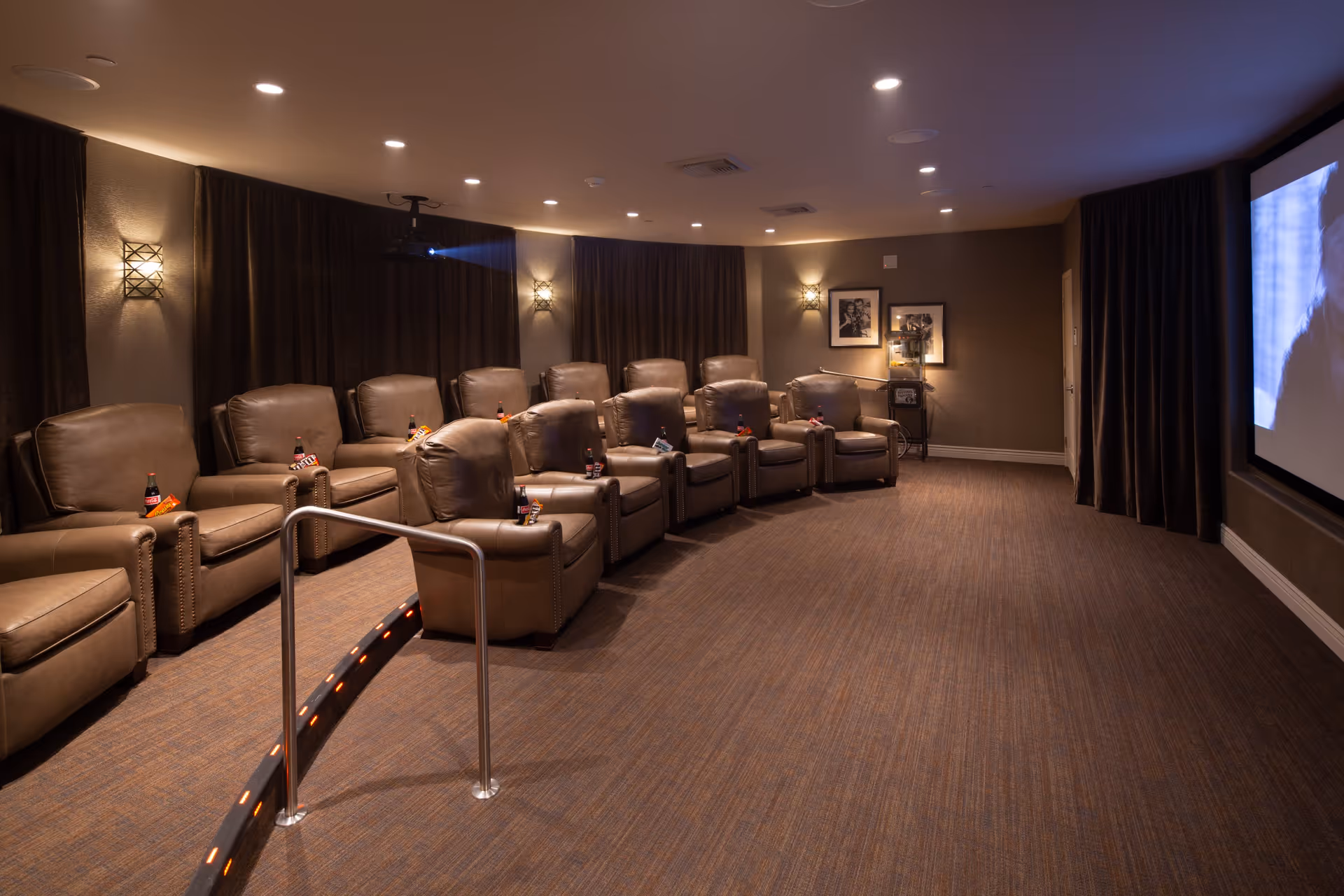 A cozy movie theater room with two rows of comfortable brown leather recliners, each with a small table holding a soda bottle and a snack. The room is dimly lit with wall sconces and has a large screen on the right side showing a black and white film. A metal handrail with small lights curves along the aisle between the rows of seats.
