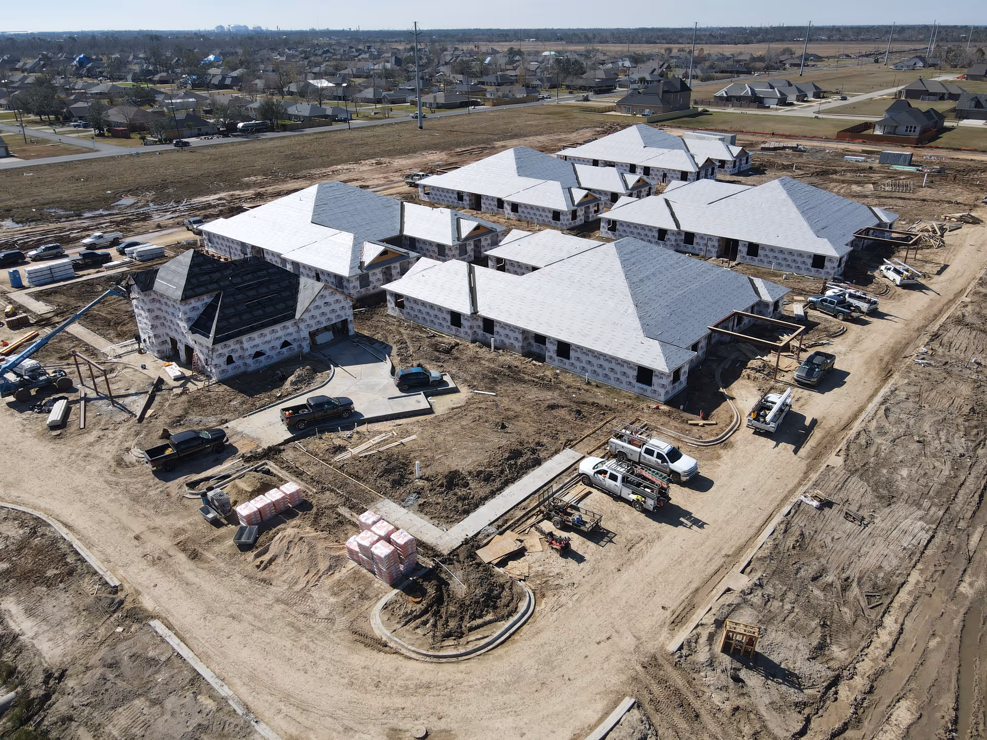 Aerial view of a construction site showing multiple buildings in progress with white roofing and wrapped exterior walls. Several trucks and construction materials are scattered around the site. The surrounding area includes residential houses and open land.