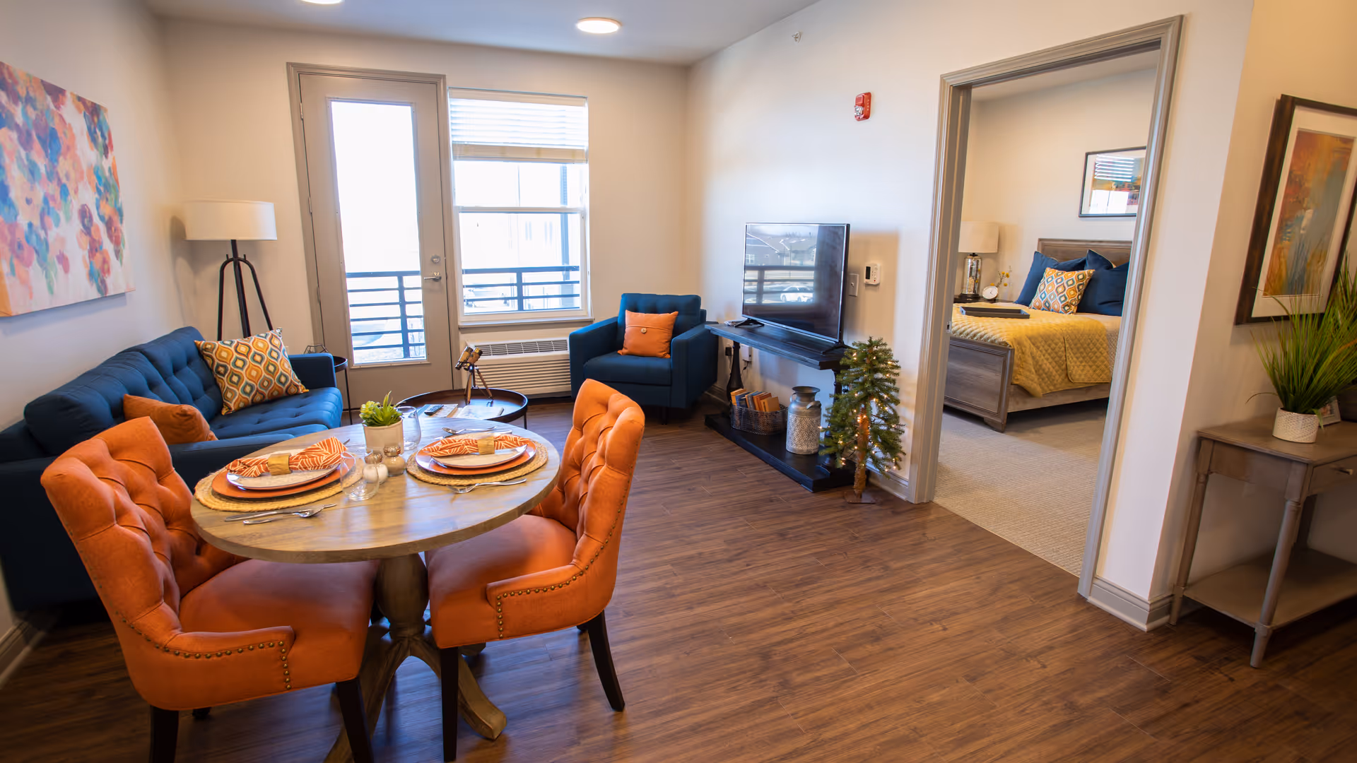 A cozy living room and dining area in a senior living facility with a round wooden dining table set for two with orange cushioned chairs. The living room has a blue sofa with patterned pillows, a matching blue armchair, a flat-screen TV on a black stand, and a small decorative plant. A doorway leads to a bedroom with a bed covered in a yellow quilt and blue pillows. The room has wooden flooring and is well-lit with natural light from a window and a glass door.