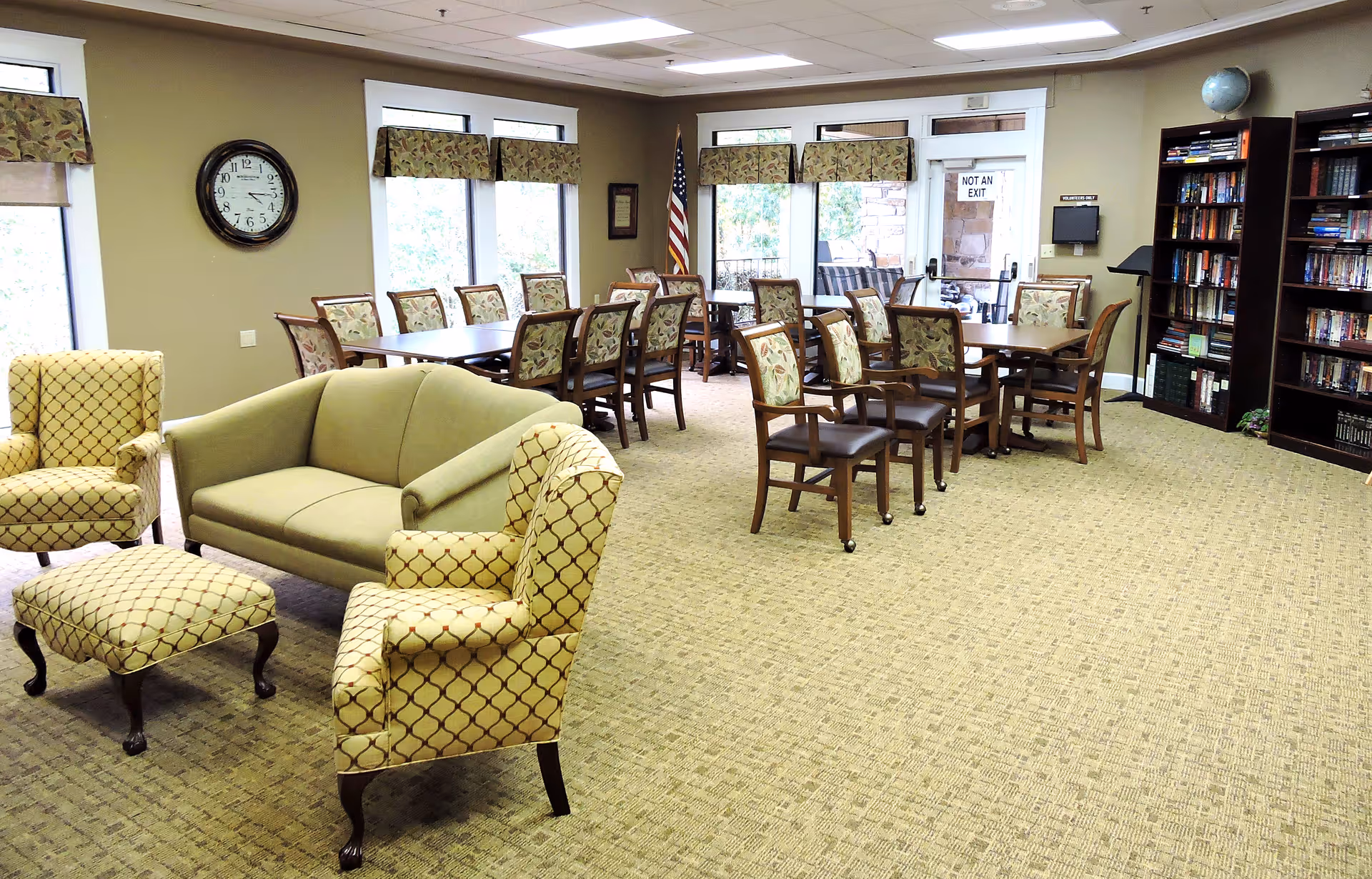 Bright communal lounge and dining area with upholstered chairs and sofas, several dining tables and chairs, and bookshelves.