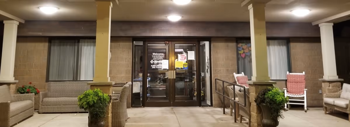 Entrance area of Crosstown Square Senior Living facility at night, featuring double glass doors with welcome signs, surrounded by brick walls and windows with curtains. The porch area is furnished with wicker sofas, chairs, rocking chairs with red cushions, and potted plants, illuminated by ceiling lights.