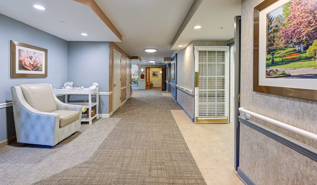 A well-lit hallway in a senior living facility with carpeted flooring and handrails along the walls. On the left side, there is a beige armchair and a small white table with stuffed animals on it. The walls are decorated with framed pictures, including one of baby feet and another of a park scene. The hallway extends into the distance with doors and additional artwork visible.