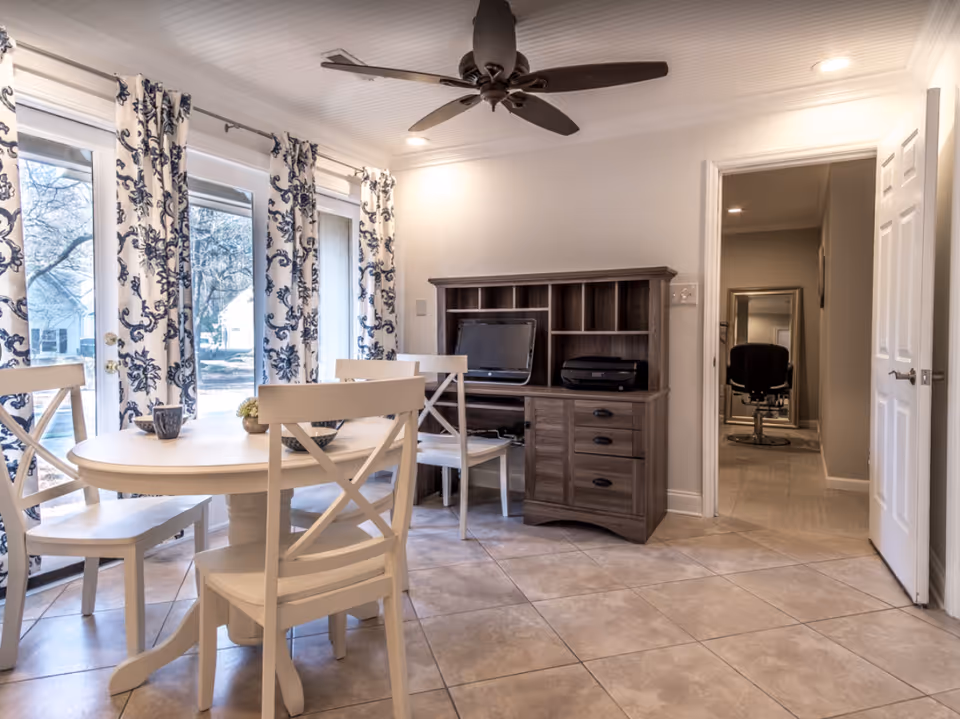 Bright dining area with a round white table and chairs by large glass doors with patterned curtains, a wooden desk with a computer, and tiled flooring.