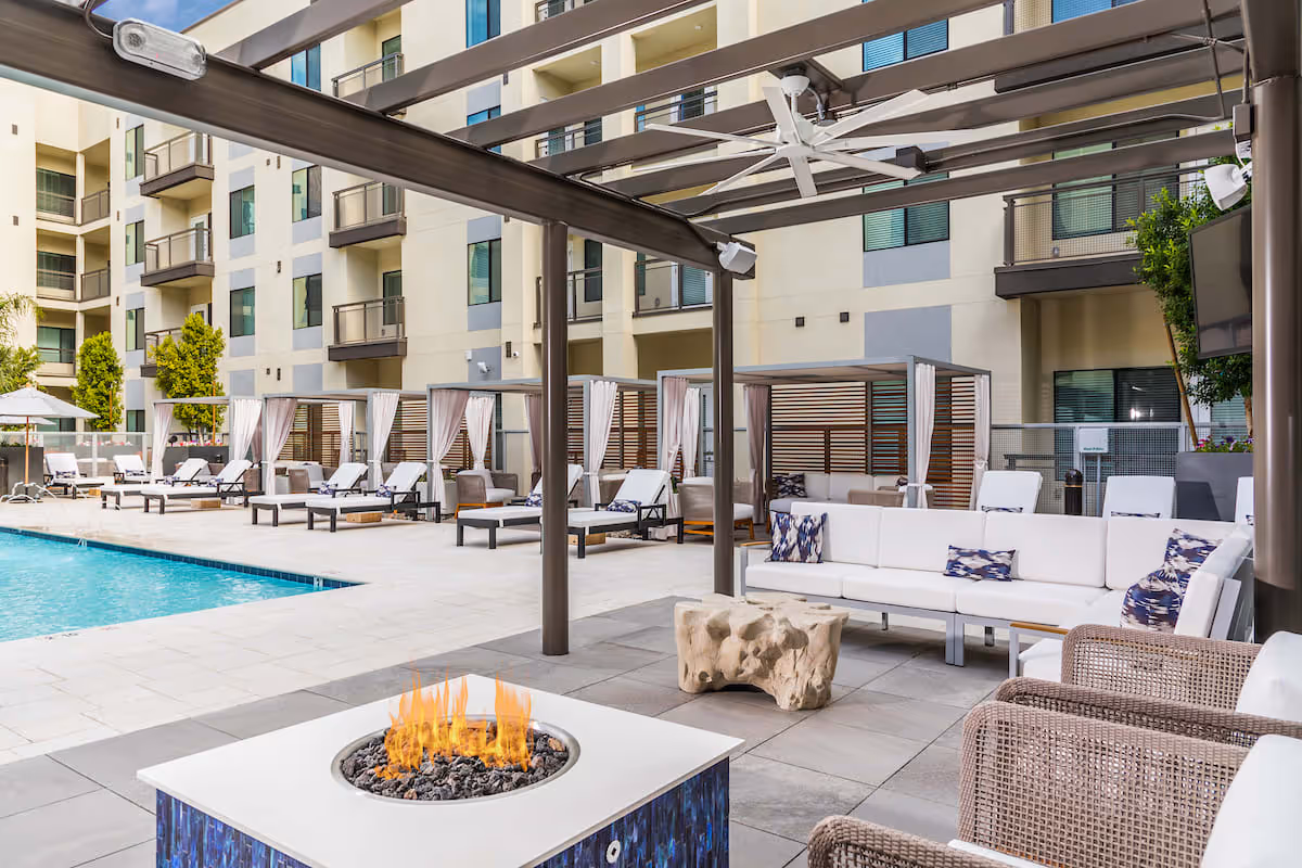 Modern outdoor poolside lounge area with cabanas, loungers, seating, and a fire pit in front of a multi-story building.