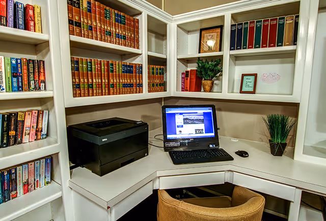 A corner office or study area with white built-in shelves filled with books, a black Dell printer, a laptop on the desk displaying a website, a computer mouse, and two small potted plants. There is a tan upholstered chair in front of the desk.