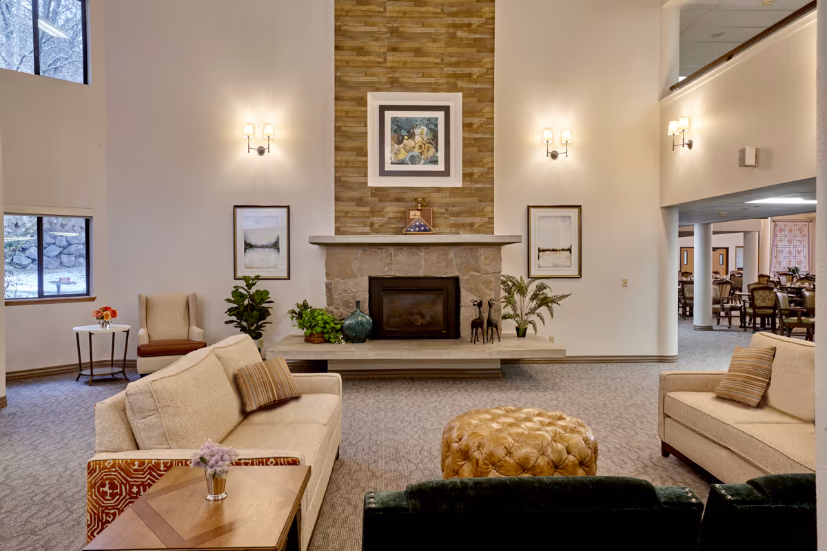 A spacious senior living facility common area featuring two beige sofas with striped pillows, a tan leather tufted ottoman, a stone fireplace with a wooden mantel, and decorative plants and artwork on the walls. The room has large windows letting in natural light and an adjacent dining area visible in the background.