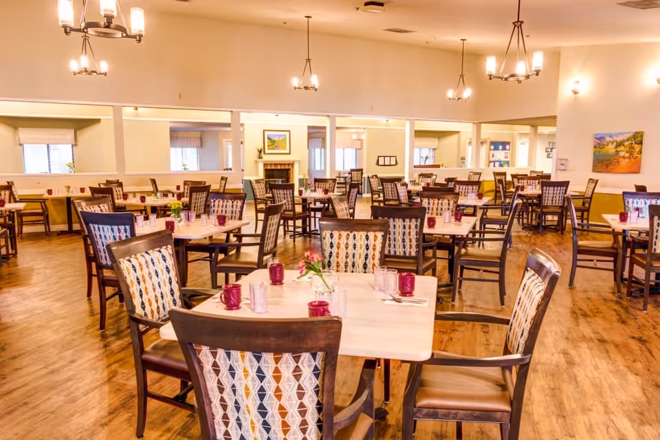 A spacious dining room with multiple square tables and wooden chairs featuring patterned backs. Each table is set with red cups, glasses, and silverware, and some have small flower arrangements. The room has wooden flooring, warm lighting from chandeliers and wall sconces, and a fireplace in the background with framed artwork above it.