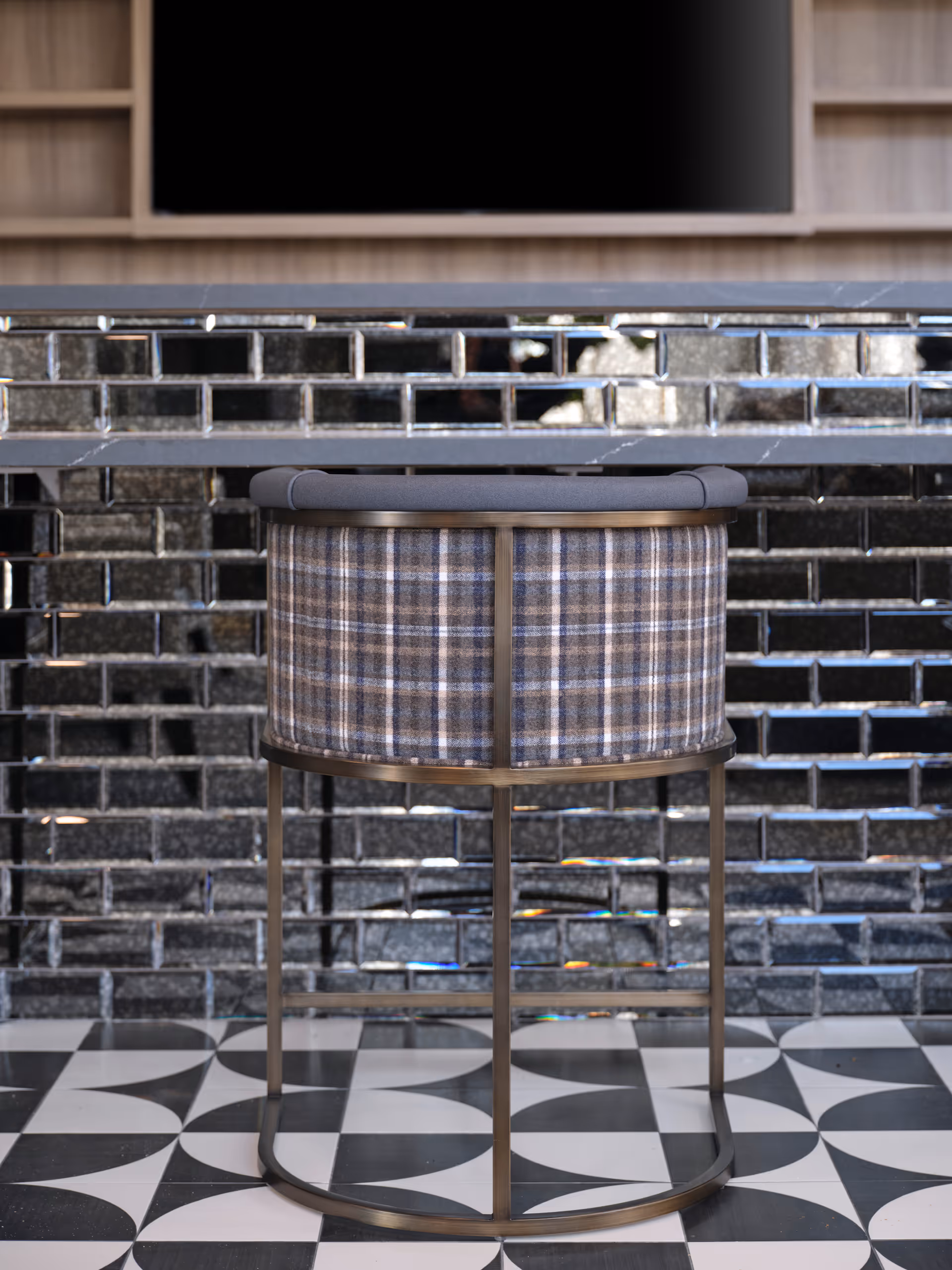 A modern chair with a metal frame and plaid fabric backrest positioned in front of a black tiled wall with a reflective surface. The floor features a black and white geometric patterned tile design. A wooden shelving unit and a large flat-screen TV are visible in the background.