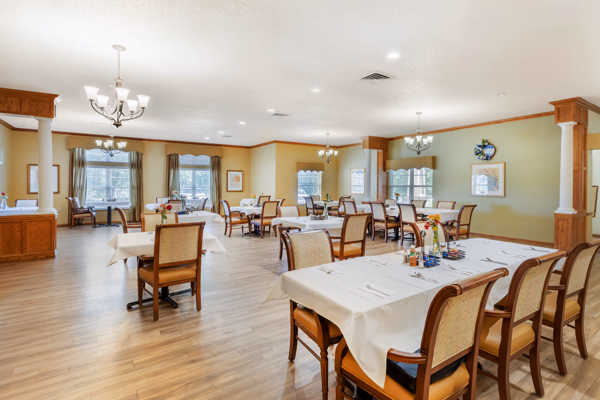 A spacious dining room with multiple tables covered in white tablecloths, each set with silverware and small flower vases. The room features wooden flooring, large windows with curtains, and decorative columns with wooden accents. Several chandeliers hang from the ceiling providing warm lighting.