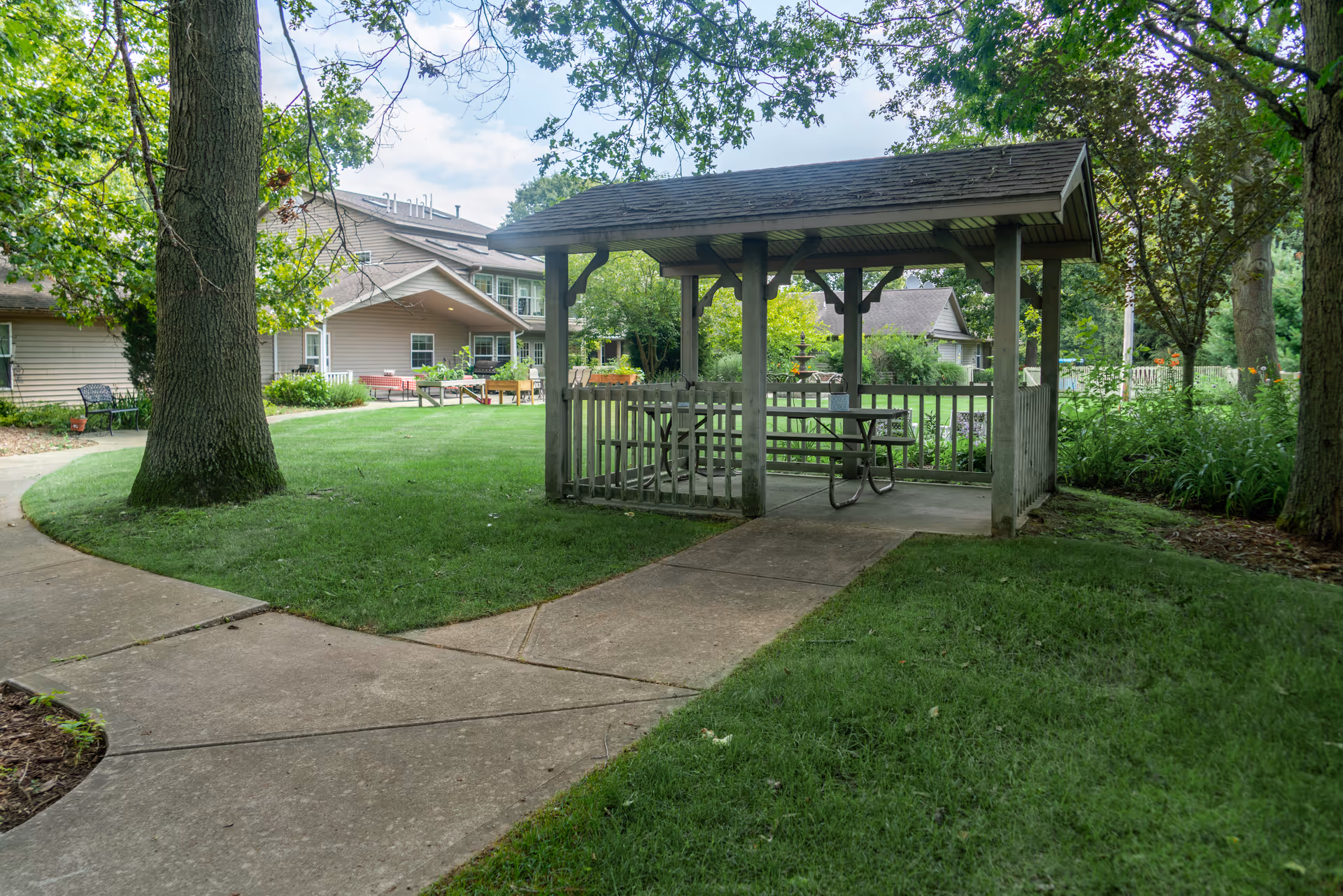 A wooden gazebo with a picnic table sits beside concrete walkways in a grassy courtyard in front of a multi-story residential building.