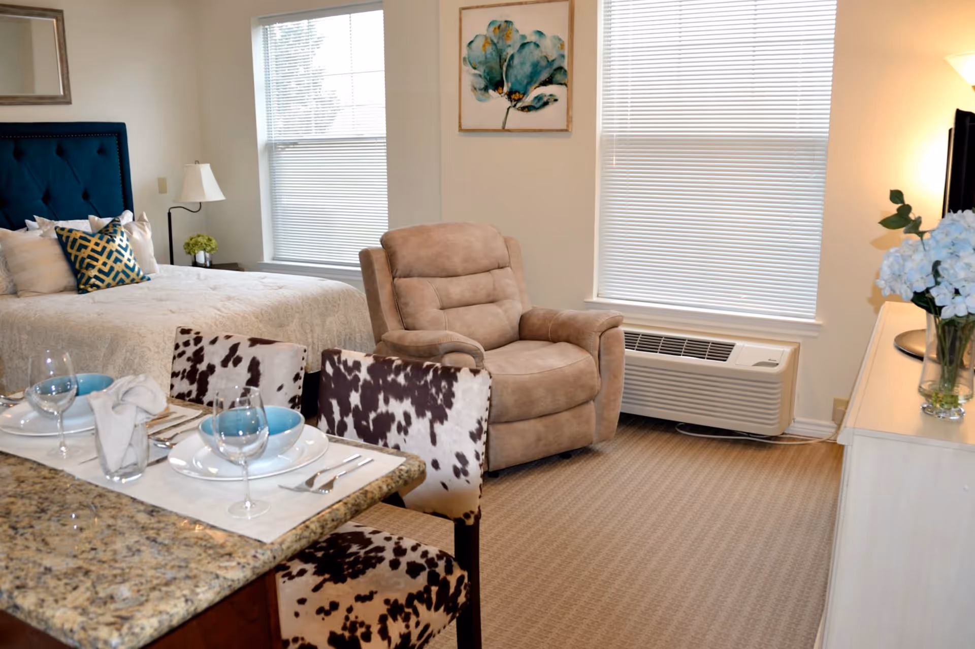 A cozy senior living facility room featuring a bed with decorative pillows, a beige recliner chair, a dining area with a granite countertop and two cowhide-patterned chairs set with plates, bowls, glasses, and silverware. The room has two windows with blinds, a white dresser with a vase of blue flowers, and a wall-mounted TV.