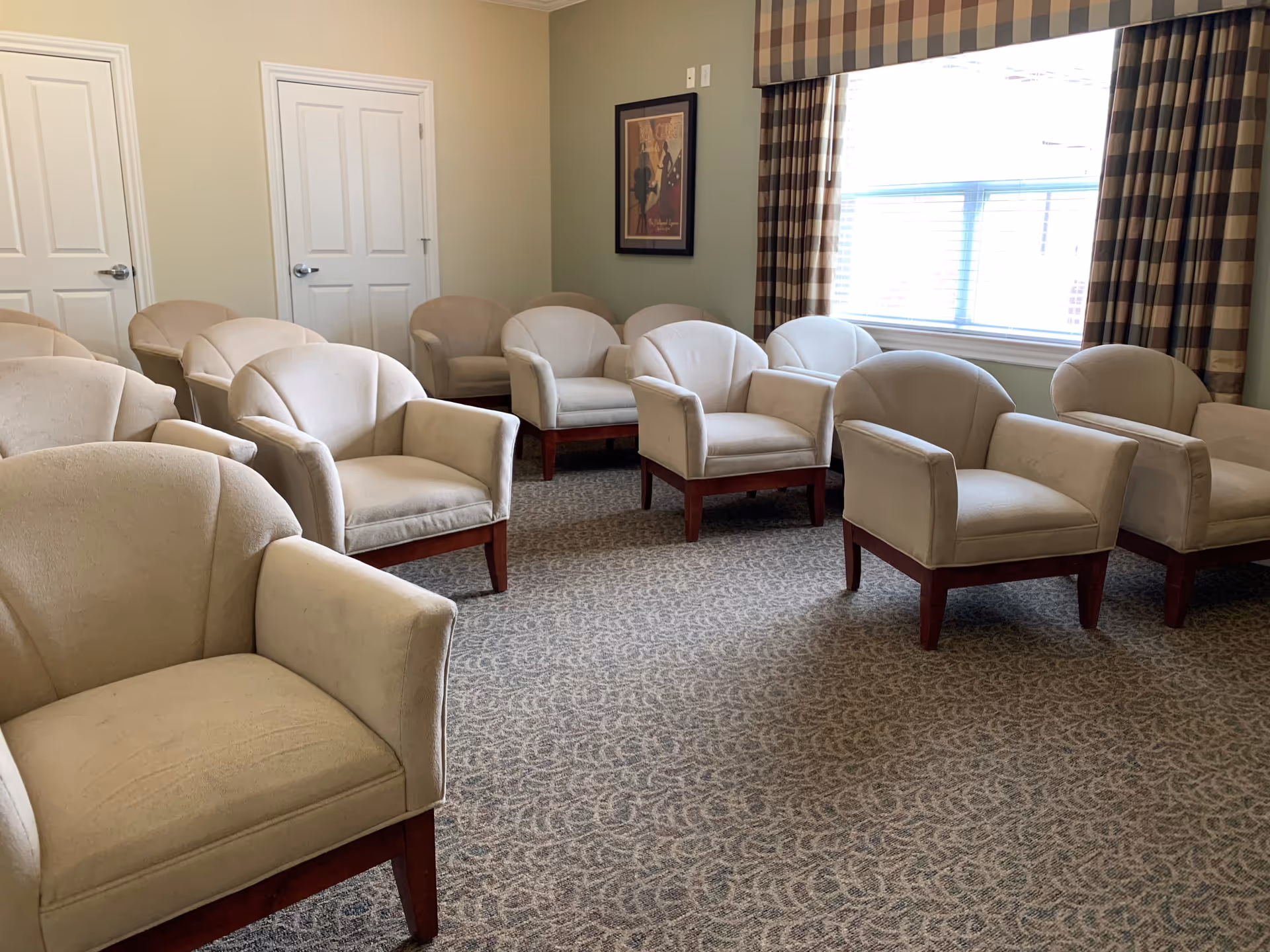 A room with multiple beige upholstered armchairs arranged in rows on a patterned carpet. There is a large window with checkered curtains on the right side, two closed white doors on the left wall, and a framed picture hanging on the wall between the doors and the window.